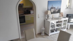 A dining area with a round table and four chairs, a white cabinet with shelves, a painting on the wall, and a kitchen visible through an archway.