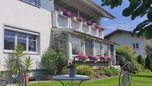 A two-story house with a balcony adorned with purple flowers, a lawn, and two metal chairs.