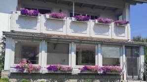 A two-story building with balconies adorned with purple flowers, a stone base, and a grassy front yard.