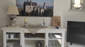 A white cabinet with shelves containing plates, cups, and a teapot. A framed picture of a castle is on the wall above the cabinet.