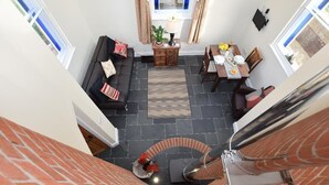Ein Raum mit hohen Decken, einer Wendeltreppe, einem Essbereich mit gedecktem Tisch für zwei Personen und einer Sitzecke mit einer Couch und Kissen.