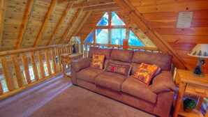 Ein gemütliches Blockhaus-Interieur mit Holzbalkendecke, einem braunen Sofa mit Kissen, einem kleinen Tisch mit Lampe und einem Fenster mit Blick auf Bäume.
