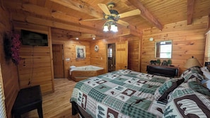 Una stanza in stile baita con un letto, una ventola a soffitto in legno, un bagno con una vasca e un pavimento in legno.