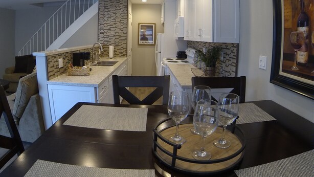 A dining area with a dark wooden table, four chairs, and a wine rack with glasses. A staircase and kitchen are visible in the background.