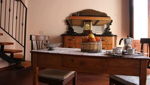 Una cocina con una mesa de madera, una cesta de frutas, una cafetera y una silla.
