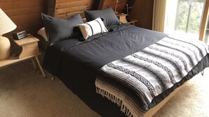 Une chambre avec un plafond en bois, un lit recouvert d’une literie sombre, une table de chevet avec une lampe et une fenêtre donnant sur des arbres.