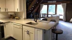 Une cuisine avec des meubles blancs, un lave-vaisselle et un tabouret de bar. Un escalier mène à une mezzanine offrant une vue sur les arbres.