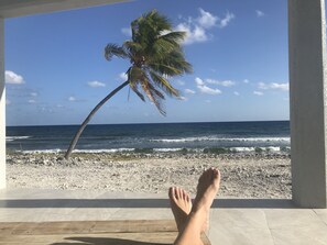 Vista de playa con una palmera, olas del océano y los pies de una persona sobre un suelo de baldosas.