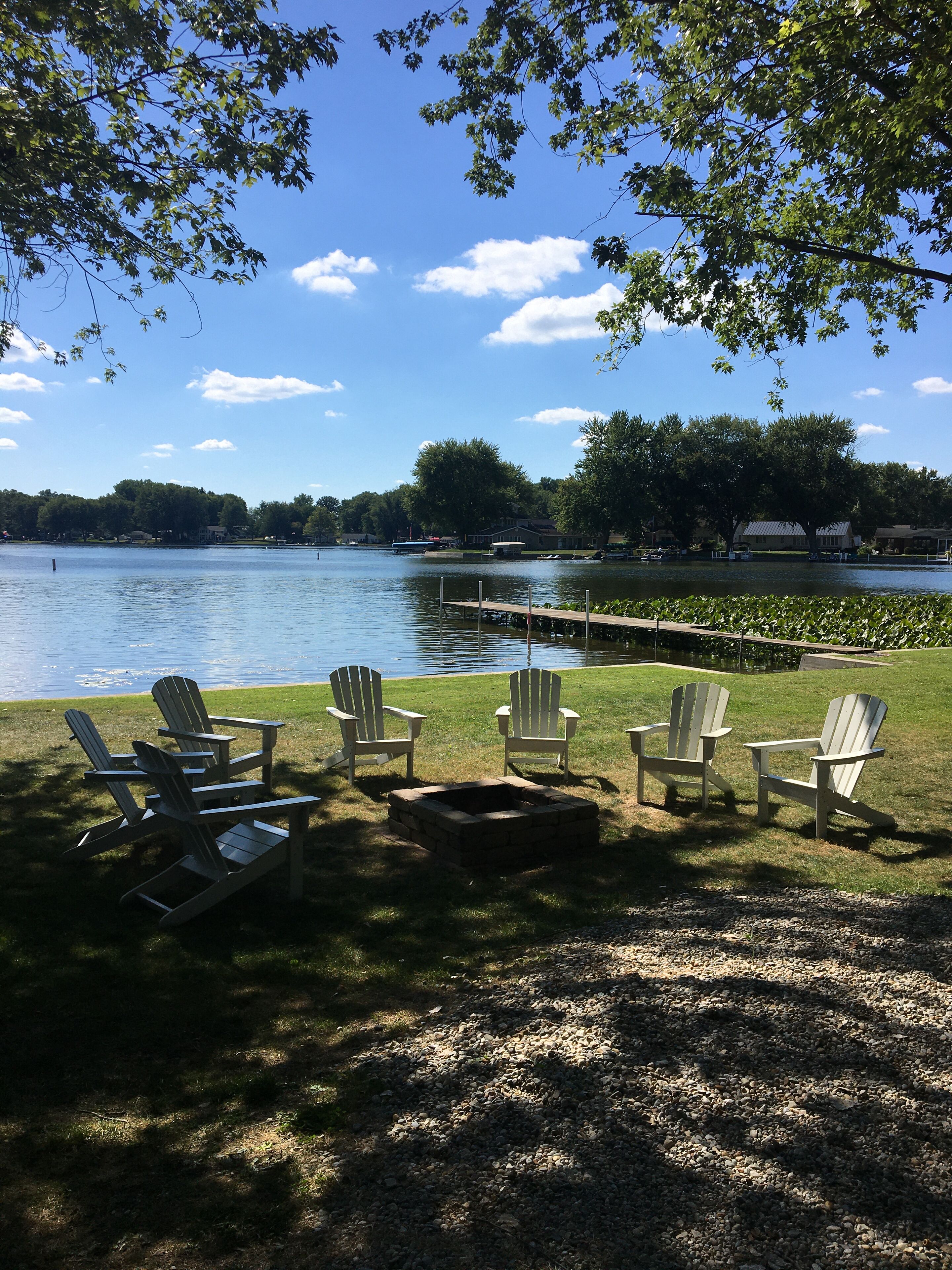 A lakeside setting with Adirondack chairs and a fire pit, surrounded by trees and a clear sky.