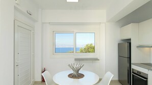 Eine moderne Küche mit weißen Schränken, einem Edelstahl-Kühlschrank und einem Backofen. Ein Esstisch mit weißen Stühlen und einem dekorativen Metall-Arrangement steht dort. Durch ein Fenster blickt man auf das Meer und Bäume.