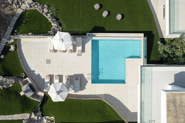 An aerial view of a modern outdoor pool area with a rectangular pool, surrounded by a stone border, and furnished with white umbrellas and lounge chairs.