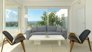 Ein modernes Wohnzimmer mit einem grauen Sofa, zwei Holzstühlen, einem weißen Couchtisch und einem großen Fenster mit Blick auf einen Pool und Grünflächen.