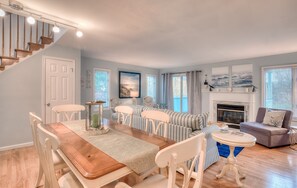 A spacious living room with a wooden dining table, striped sofas, a fireplace, and large windows.