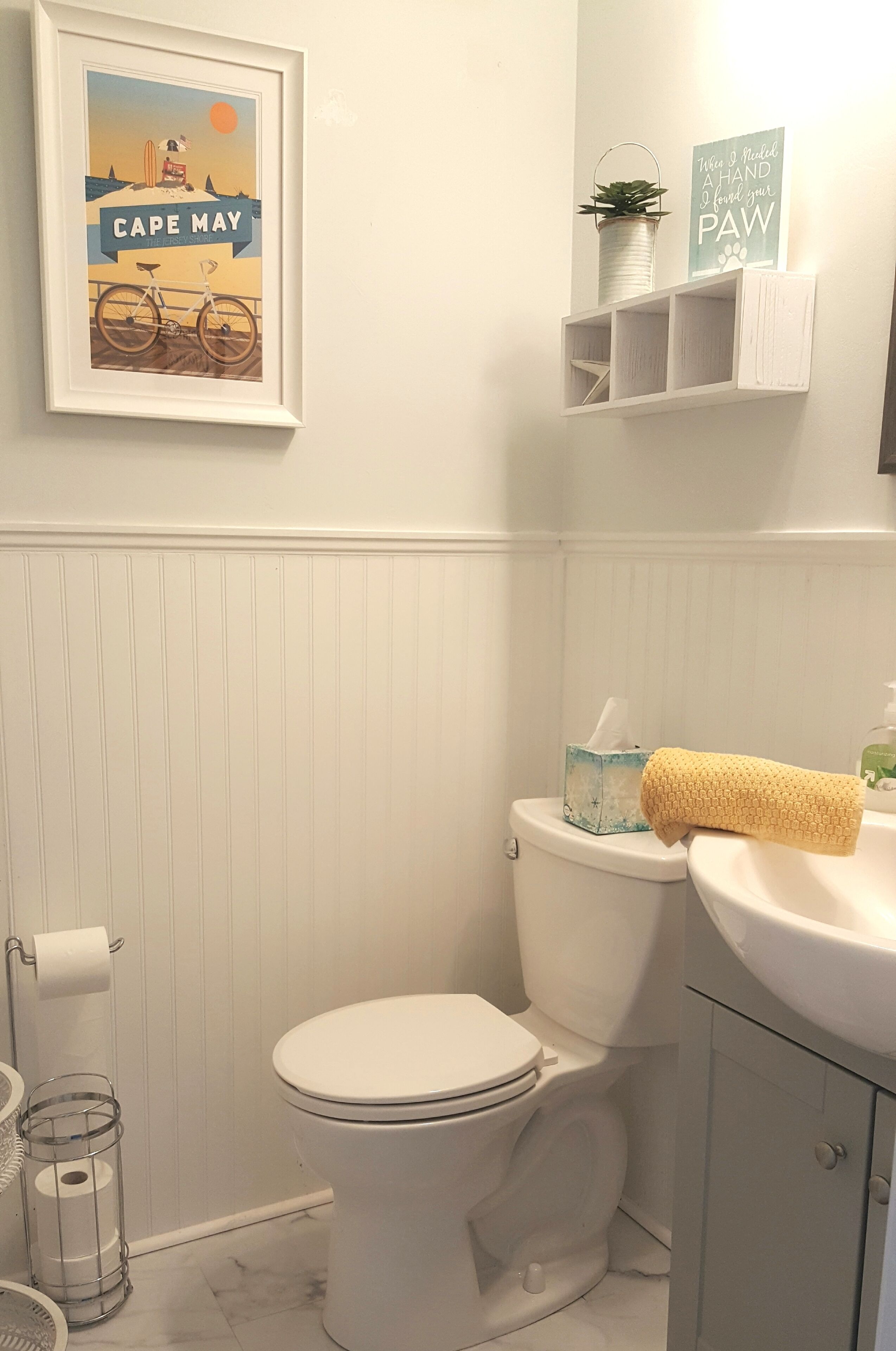 A bathroom with a toilet, a sink, a framed picture of Cape May, and a small shelf with a plant and a sign.