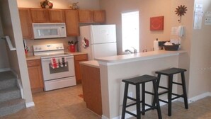 A kitchen with wooden cabinets, a white refrigerator, a microwave, and a stove. There are two black bar stools and a window with blinds. - Perfect Family Vacation Spot close to major Orlando attractions! (Kissimmee)