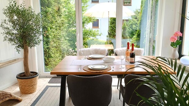 A dining table set for two with a potted plant, wine bottles, and a view of greenery outside.