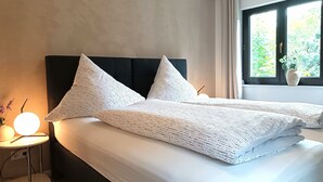 A neatly made bed with a white sheet and two pillows, a bedside table with a lamp, and a window with a view of greenery.
