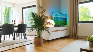 A modern living room with a TV, a dining area with chairs, a large plant, and a view of greenery outside.