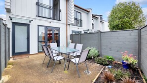 Een modern huis met twee verdiepingen, met een patio voorzien van een glazen tafel en stoelen, omgeven door een houten hek en een tuin met verschillende planten.