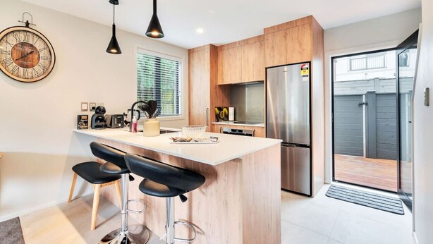 Een moderne keuken met een centraal eiland, barkrukken, een grote klok en een schuifdeur naar buiten.