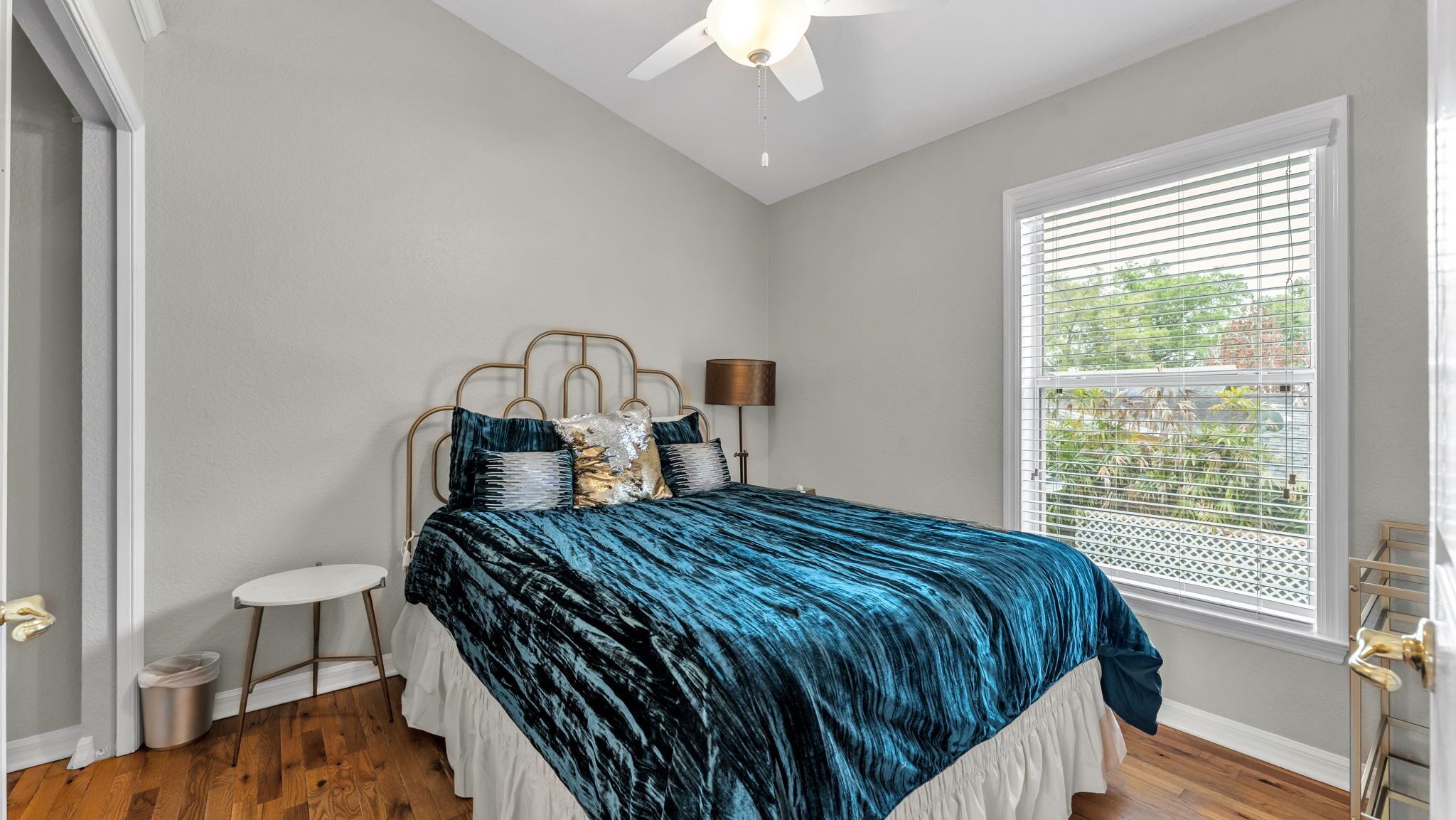 Quarto com cama, ventilador de teto, janela com persianas e uma pequena mesa de cabeceira.