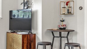 Un salon moderne avec un téléviseur à écran plat posé sur un meuble en bois, une table de salle à manger ronde avec des tabourets, et une étagère décorative agrémentée d’une horloge et de fleurs.