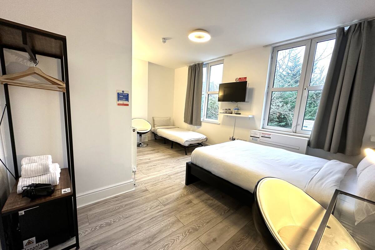A hotel room with a bed, bedside table, chair, TV, and a window with curtains.