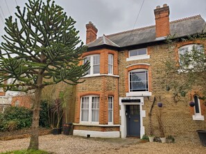 A traditional brick house with a prominent tree in the front yard, a clear sign reading "SLEEP ELICITY," and a well-maintained garden. - Sleeplicity London Heathrow (Feltham)