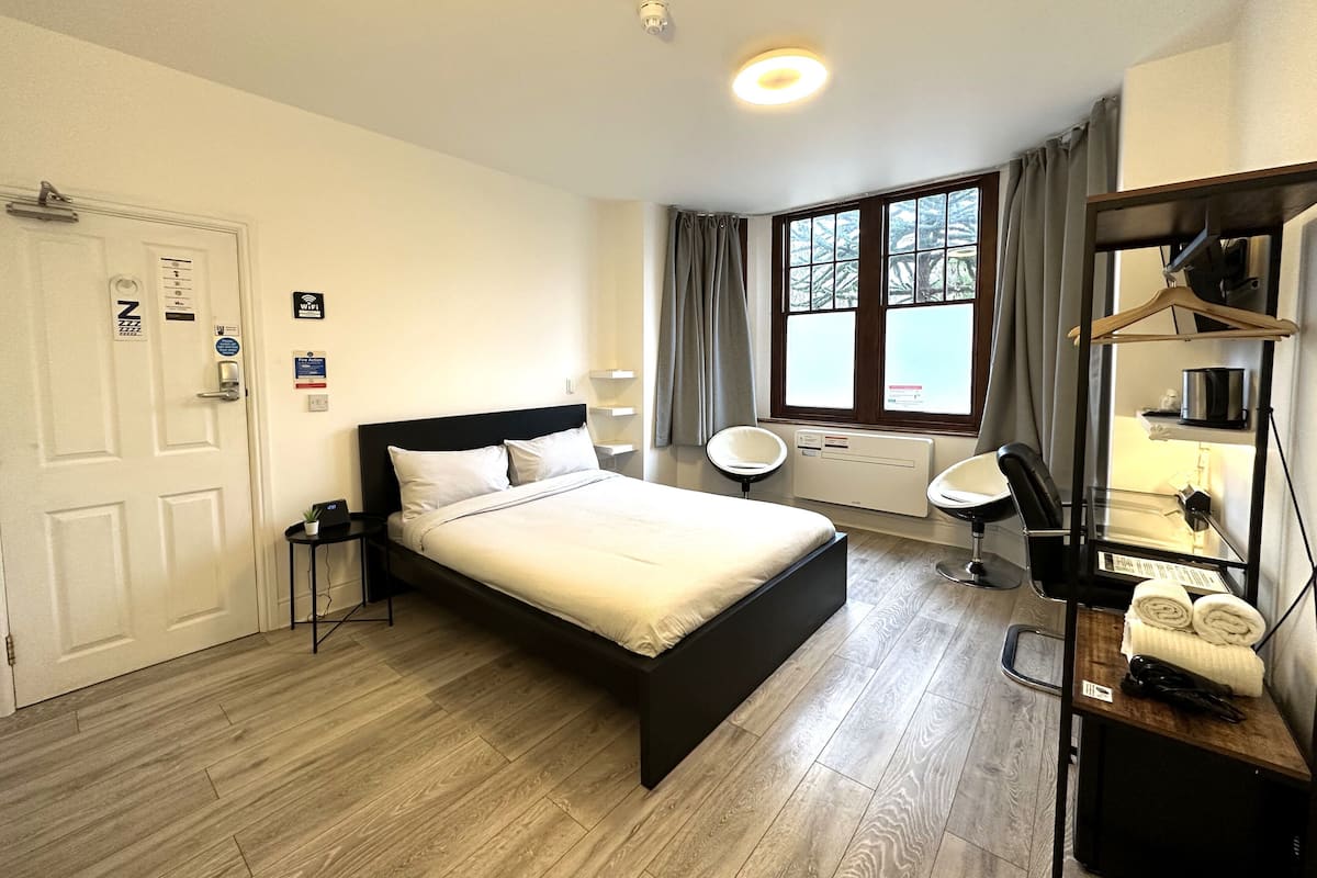 A hotel room with a bed, bedside tables, a desk with a chair, a window with curtains, and a radiator.