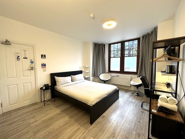 A hotel room with a bed, bedside tables, a desk with a chair, a window with curtains, and a radiator. - Sleeplicity London Heathrow (Feltham)