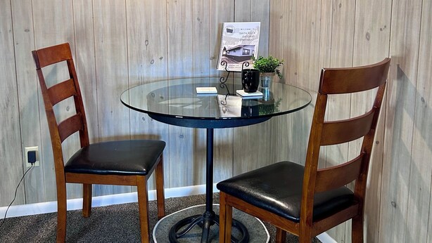 Ett litet mötesrum med ett runt bord av glas, två trästolar, en inramad välkomstskylt och en landskapsmålning på väggen.
