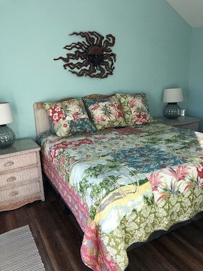 Un dormitorio con edredón estampado de flores, dos lámparas de noche, un tocador de madera y un adorno en la pared.