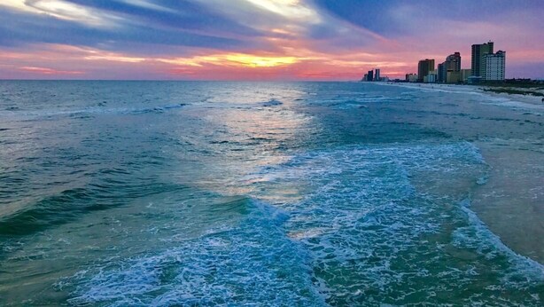 A coastal cityscape with a sunset over the ocean, featuring a beach and high-rise buildings.