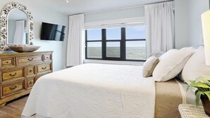 A bedroom with a large bed, a ceiling fan, a TV, a window with a view of the sea, and a wooden dresser.