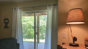 A room with a sliding glass door leading to a garden, a wooden table with a lamp, and a grey sofa. - Fishermen and Family’s Lakeside Nature Retreat  (Cape Fair)