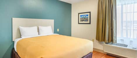 A hotel room with a bed, a window with curtains, a wall-mounted air conditioner, and a framed picture on the wall.