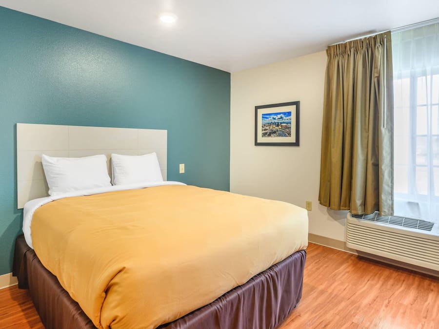 A hotel room with a bed, a window with curtains, a wall-mounted air conditioner, and a framed picture on the wall.