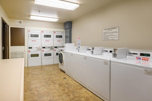 A hotel laundry facility with multiple washing machines and dryers, a sign with guest laundry guidelines, and a ventilation system on the ceiling.
