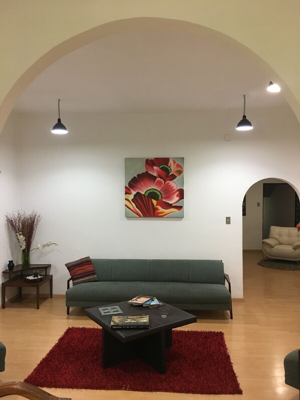 A living room with a green sofa, a coffee table, and a red rug. There is a painting of flowers on the wall and two pendant lights.