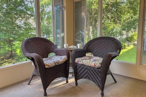 Dos sillones de mimbre con cojines estampados, una mesita con taza y platillo, y una vista de un paisaje verde a través de amplios ventanales.