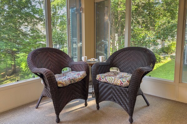 Two wicker chairs with patterned cushions, a small table with a cup and saucer, and a view of a green landscape through large windows. - Walk-In Condo With Pools in Pointe Royale - 2 Kings - Sleeps 6 (Branson)