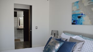A bedroom with a bed, pillows, a bedside table, a lamp, a painting, and a view into the bathroom. - Cozy Stays Cayala Apartments 3 (Guatemala)