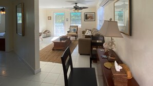 A spacious living room with a ceiling fan, a sofa, a coffee table, and a lamp. There is a door leading to another room and a window with blinds. - A garden oasis for the nature lover who likes modern comforts (Gregory Town)