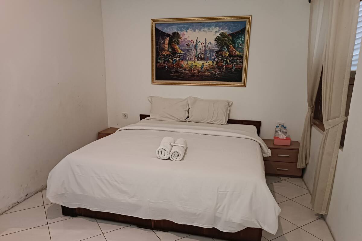A neatly made bed with white linens, a painting above it, and a small bedside table with a tissue box.