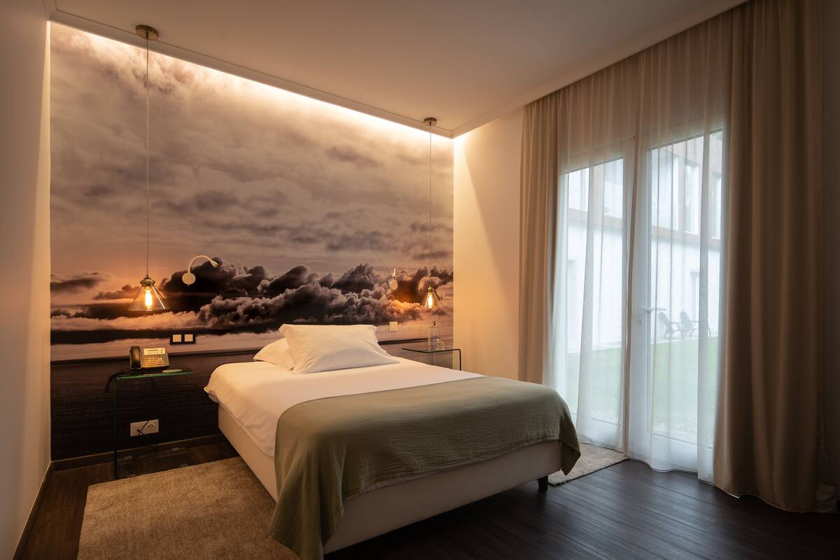 A modern bedroom with a large bed, a nightstand with a lamp, a wall mural of clouds, and a large window with curtains.