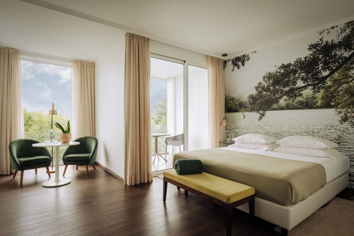 A hotel room with a bed, two chairs, a small table, a bench, and a large window with curtains.