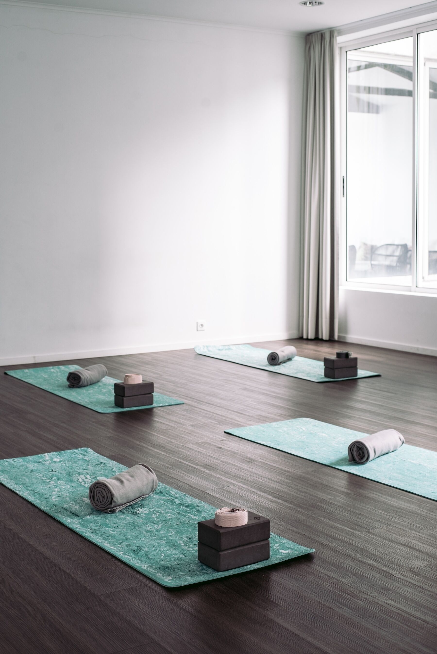 A room with wooden flooring, large windows with curtains, and yoga mats with blocks and rolled-up mats.