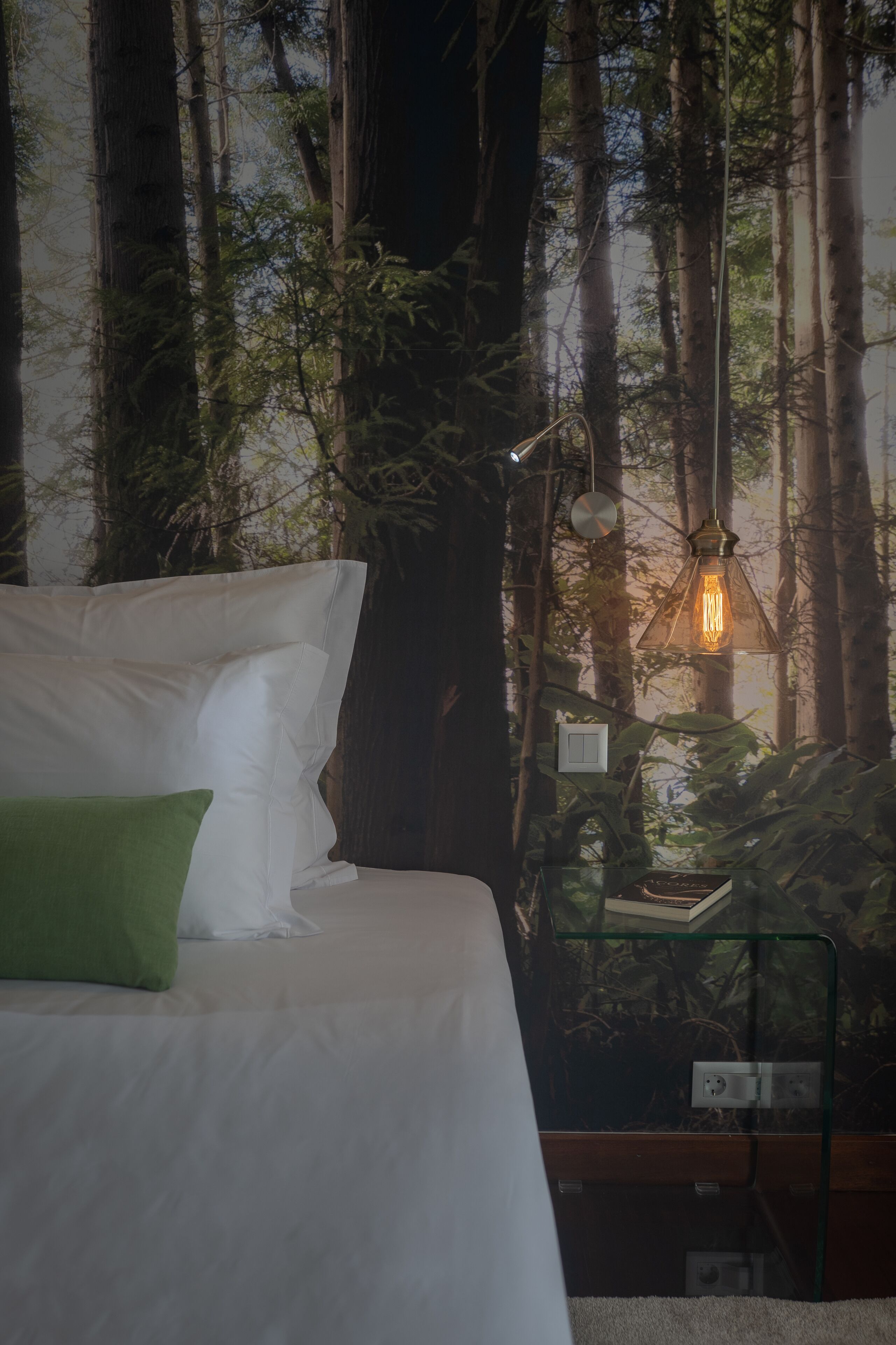 A bedroom with a bed, a green pillow, a nightstand, a lamp, and a wall with a forest mural.