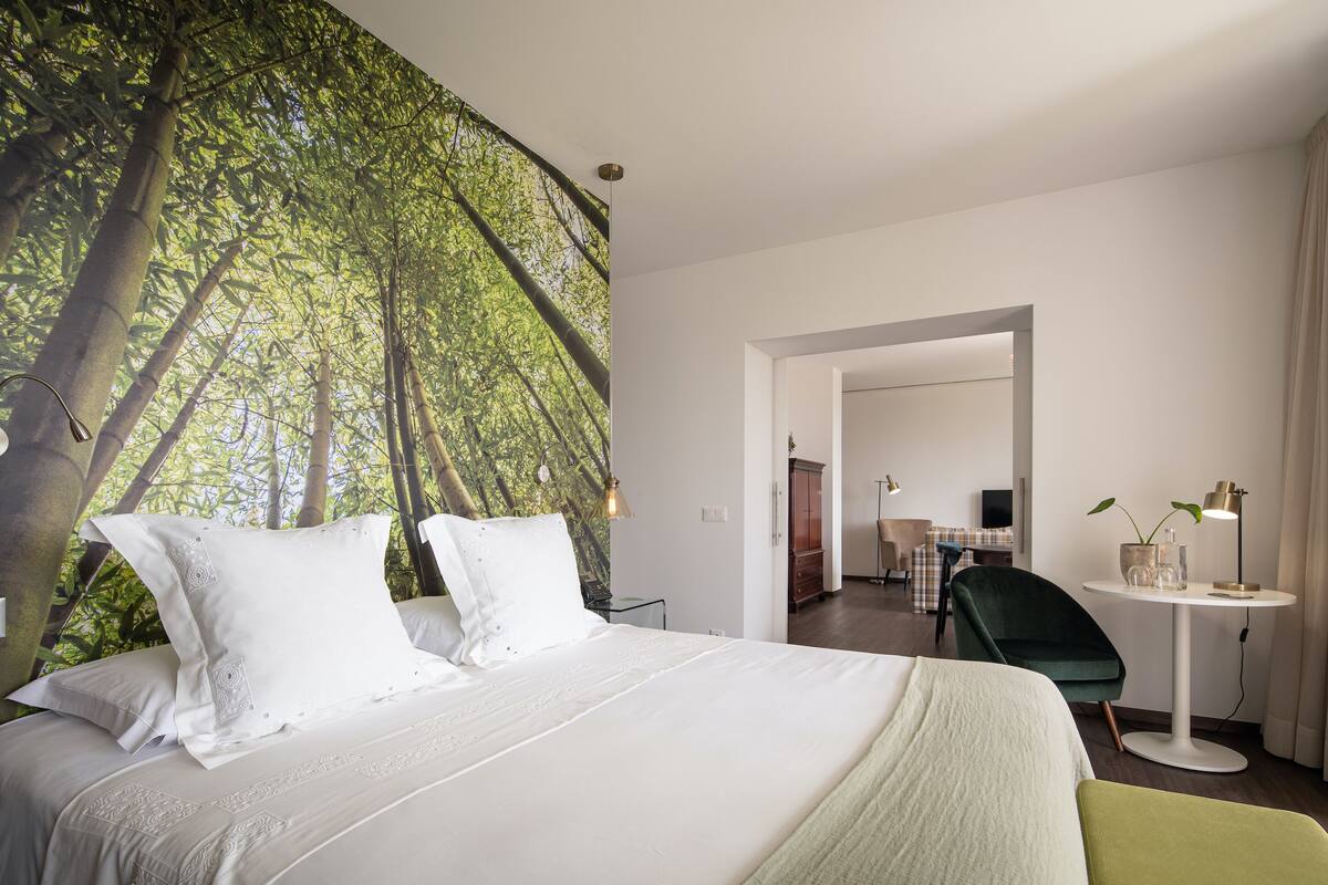 A hotel room with a large bed, a desk with a lamp, and a view of a forest through a large window.