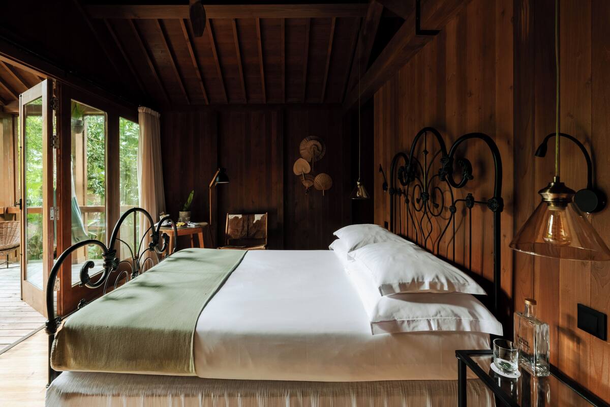 A wooden room with a bed, a ceiling fan, a table with a lamp, and a window.